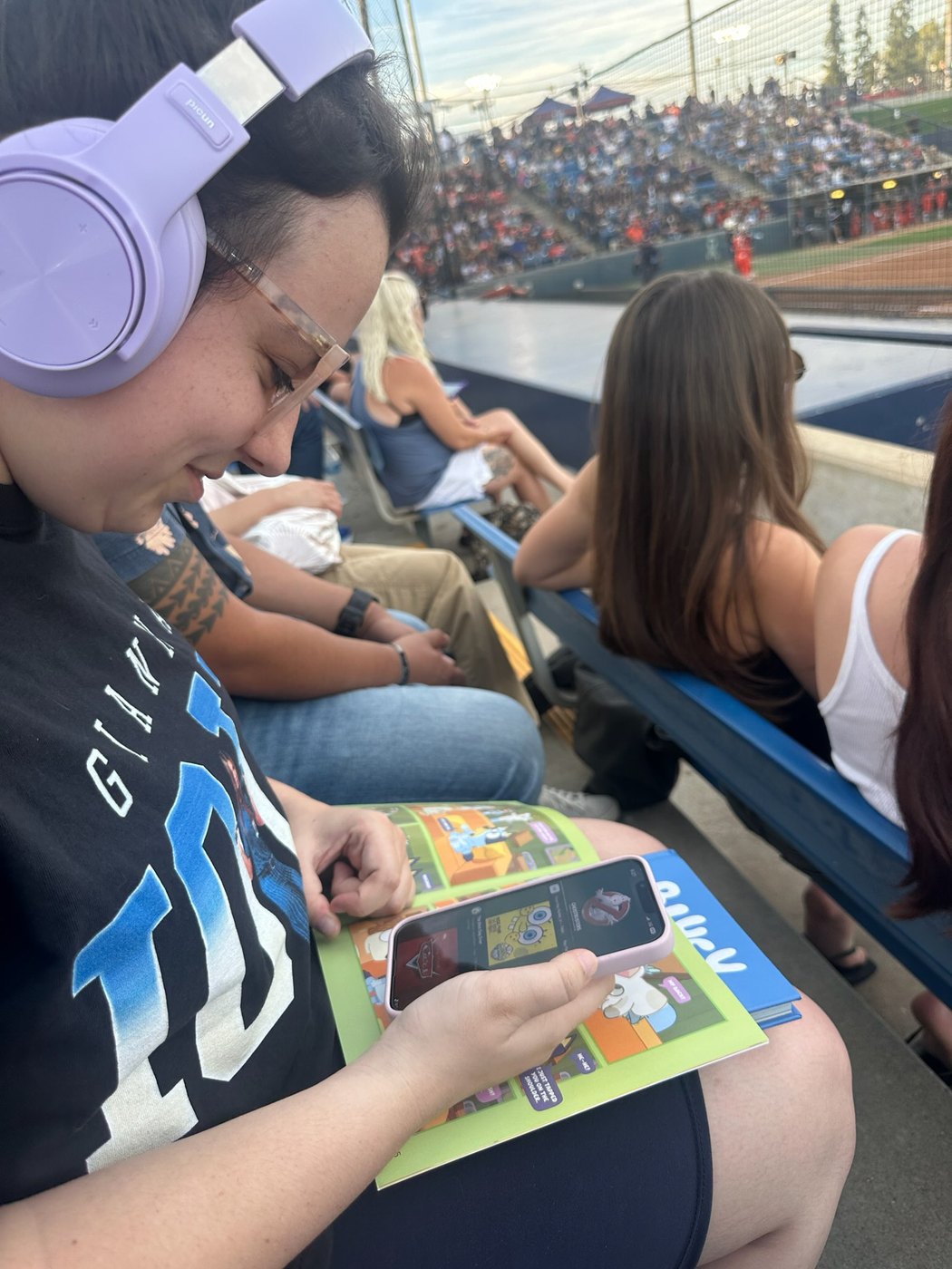 A young person at a ballgame wearing noise-reducing headphones, watching cartoons on a phone with an activity book on her lap