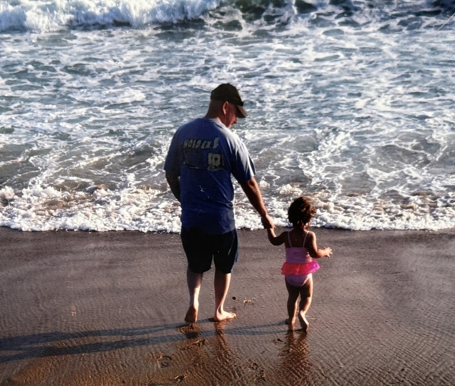 A father and his young daughter holding hands at the edge of the ocean, seen from behind